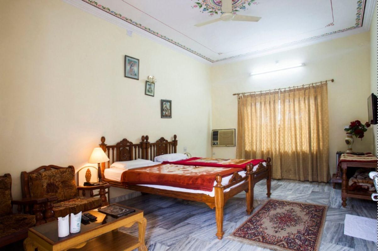 Quarto de hotel com cama, escrivaninha, cadeira, abajur, ventoinha no teto, janela com cortinas e ar condicionado fixado na parede.