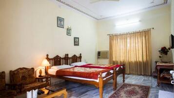 Quarto de hotel com cama, escrivaninha, cadeira, abajur, ventoinha no teto, janela com cortinas e ar condicionado fixado na parede.