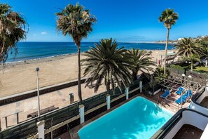 Une piscine avec vue sur la plage et l’océan, entourée de palmiers et de chaises longues.