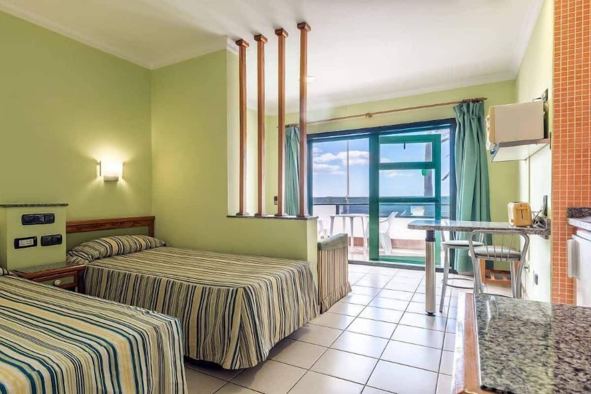 A hotel room with two beds, a small kitchenette, a balcony with a view, and a large window.