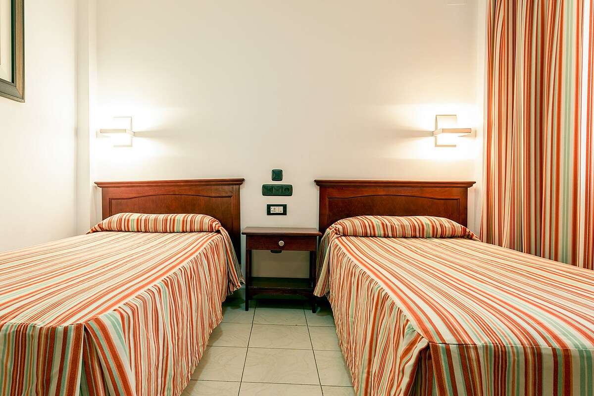 Habitación doble con dos camas individuales, una mesita de noche y una lámpara fijada a la pared.