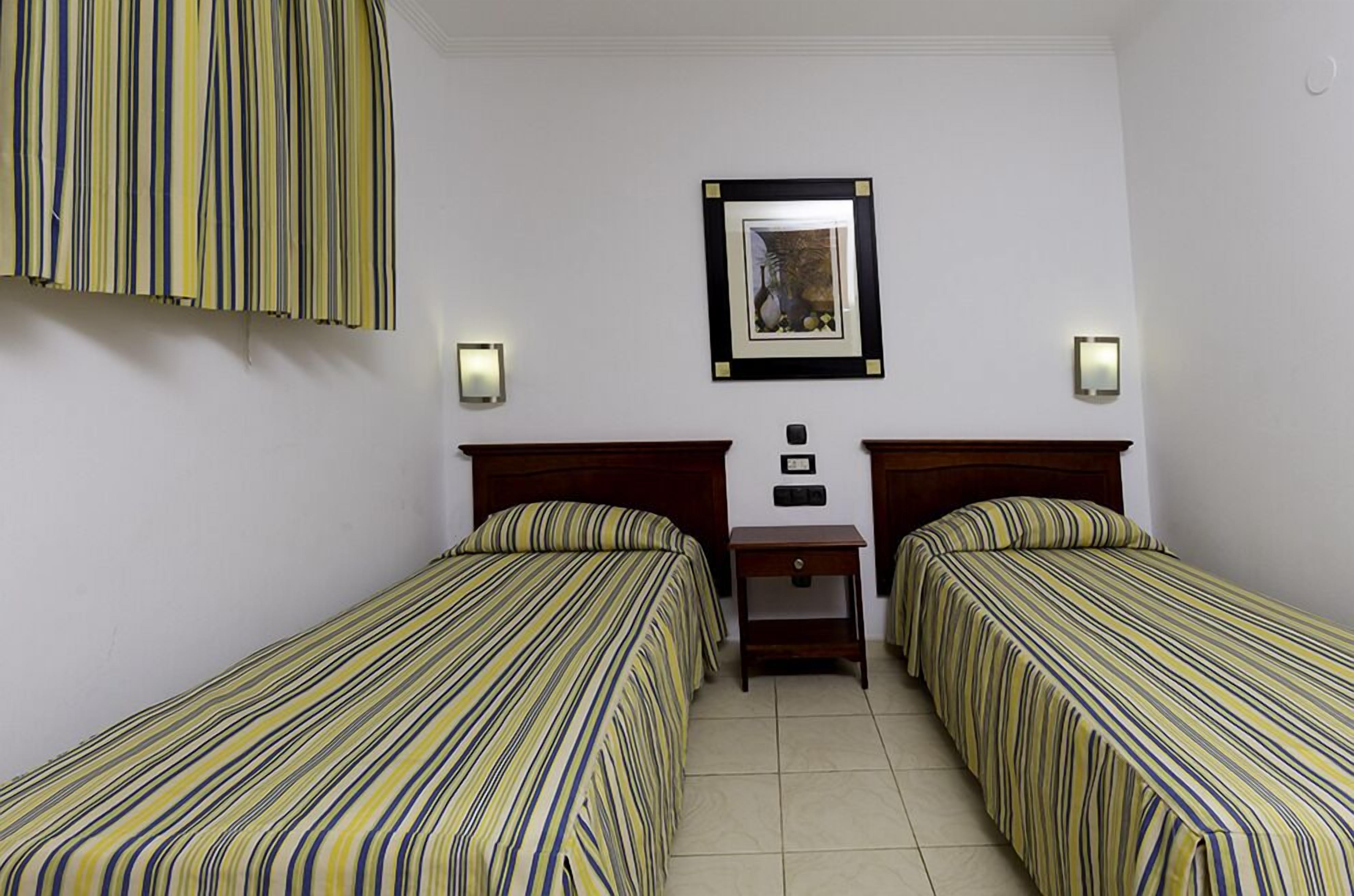A hotel room with two single beds, a small bedside table, a framed picture on the wall, and two wall-mounted lights.