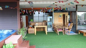 A covered outdoor area with artificial grass, wooden furniture, a hot tub, and a refrigerator. Flags are hanging from the ceiling. - Nap Inn (Ko Pha-ngan)