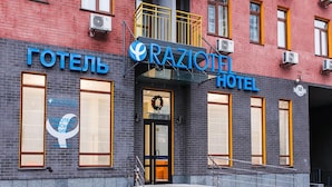 Eine Hotelansicht mit einer auffälligen Schrifttafel, auf der "RAZIOTEL HOTEL" steht, und einem Logo. Es gibt große Fenster und einen gut erkennbaren Eingang mit Stufen, die dorthin führen.