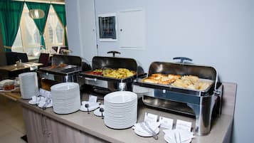 Montaje de bufé con diversos alimentos en recipientes de metal, platos apilados y utensilios para servir sobre una barra.