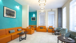 Ein modernes Wohnzimmer mit orangefarbenen Sofas, einem Couchtisch und einem grünen Sessel. Zwei Fenster mit Jalousien, eine Deckenleuchte und gerahmte Kunstwerke an den Wänden sind ebenfalls vorhanden.