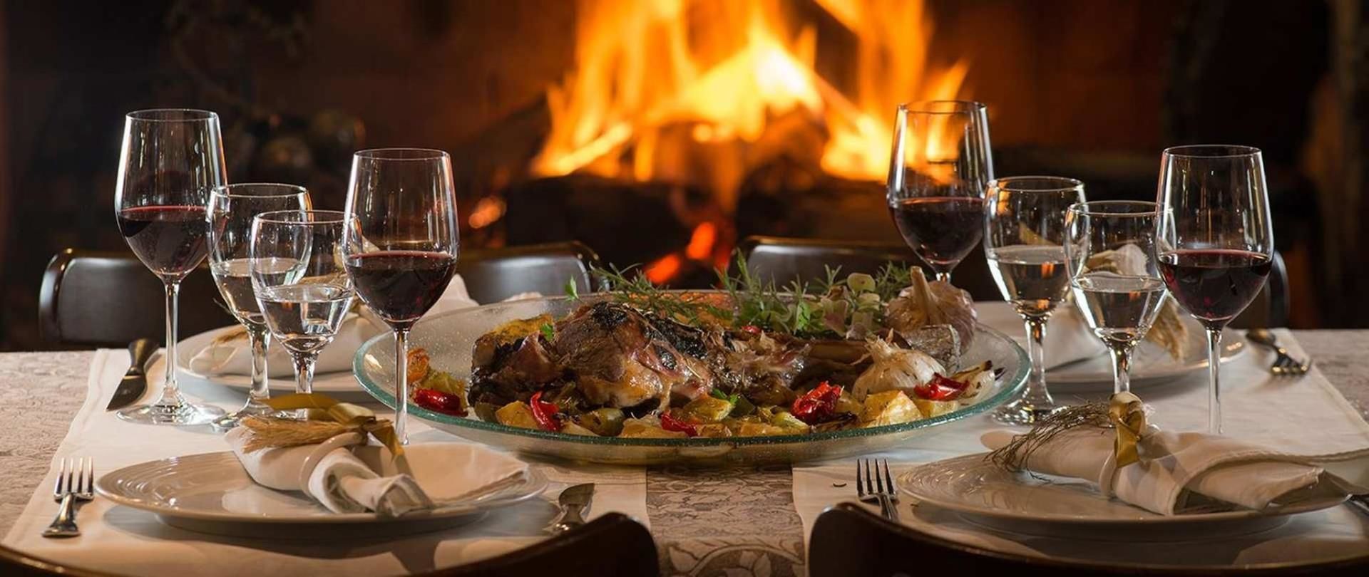 A dining table set with plates, glasses, and cutlery, with a roasted dish in the center and a fireplace in the background.