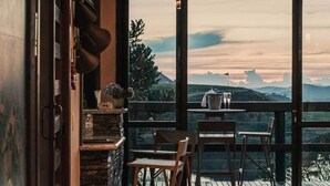 Une pièce chaleureuse avec une grande fenêtre donnant sur un paysage pittoresque, une table en bois avec des chaises et une décoration rustique agrémentée de pots suspendus.
