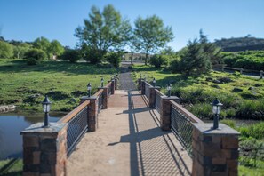 A bridge with stone railings and lanterns, leading to a pathway surrounded by greenery and a body of water.