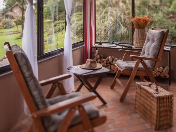 A cozy seating area with two wooden chairs and a small table, a wicker basket, and a window with a view of greenery outside.