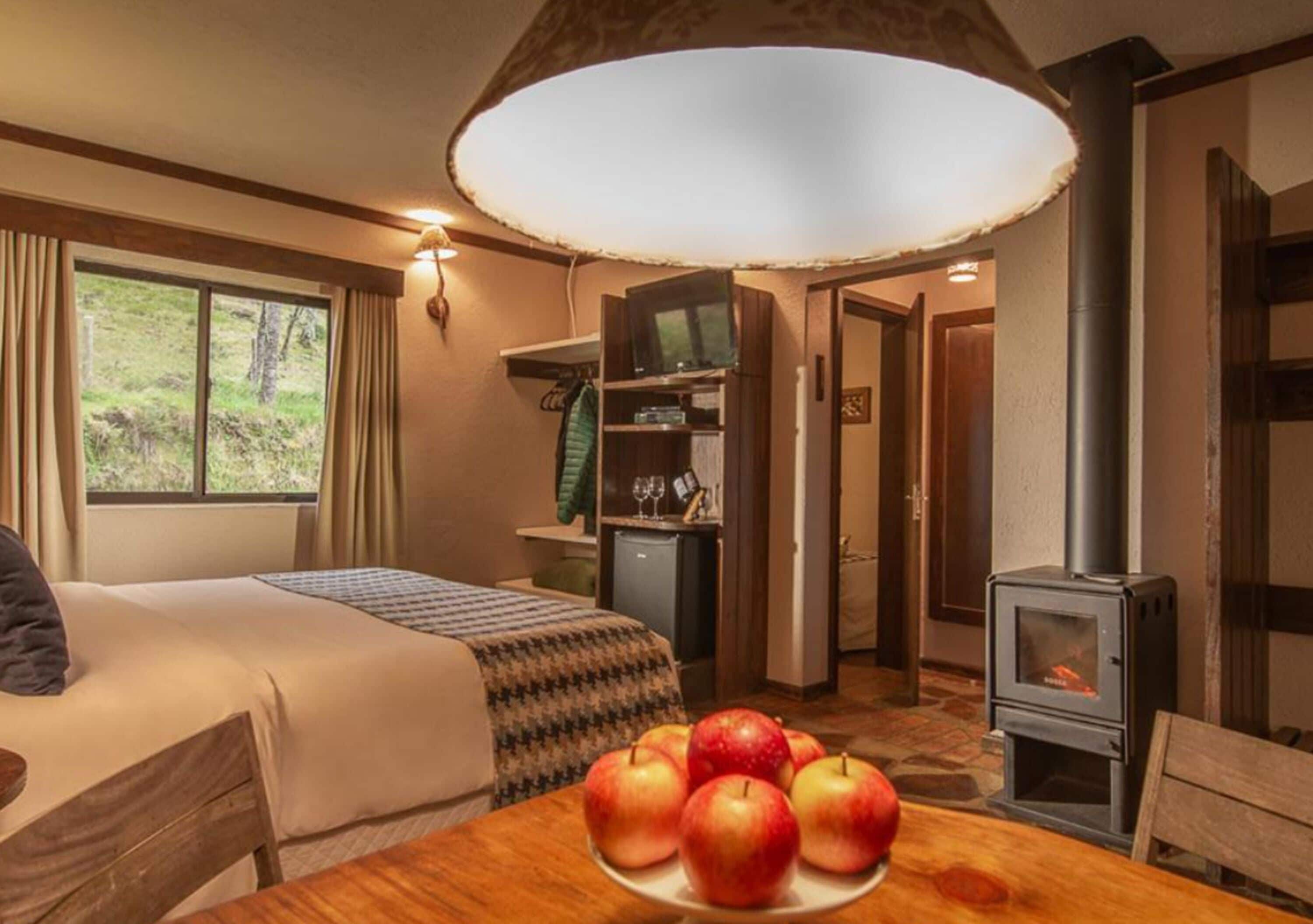 Une pièce confortable avec un lit, une table sur laquelle sont posées des pommes, une cheminée et une fenêtre avec des rideaux.