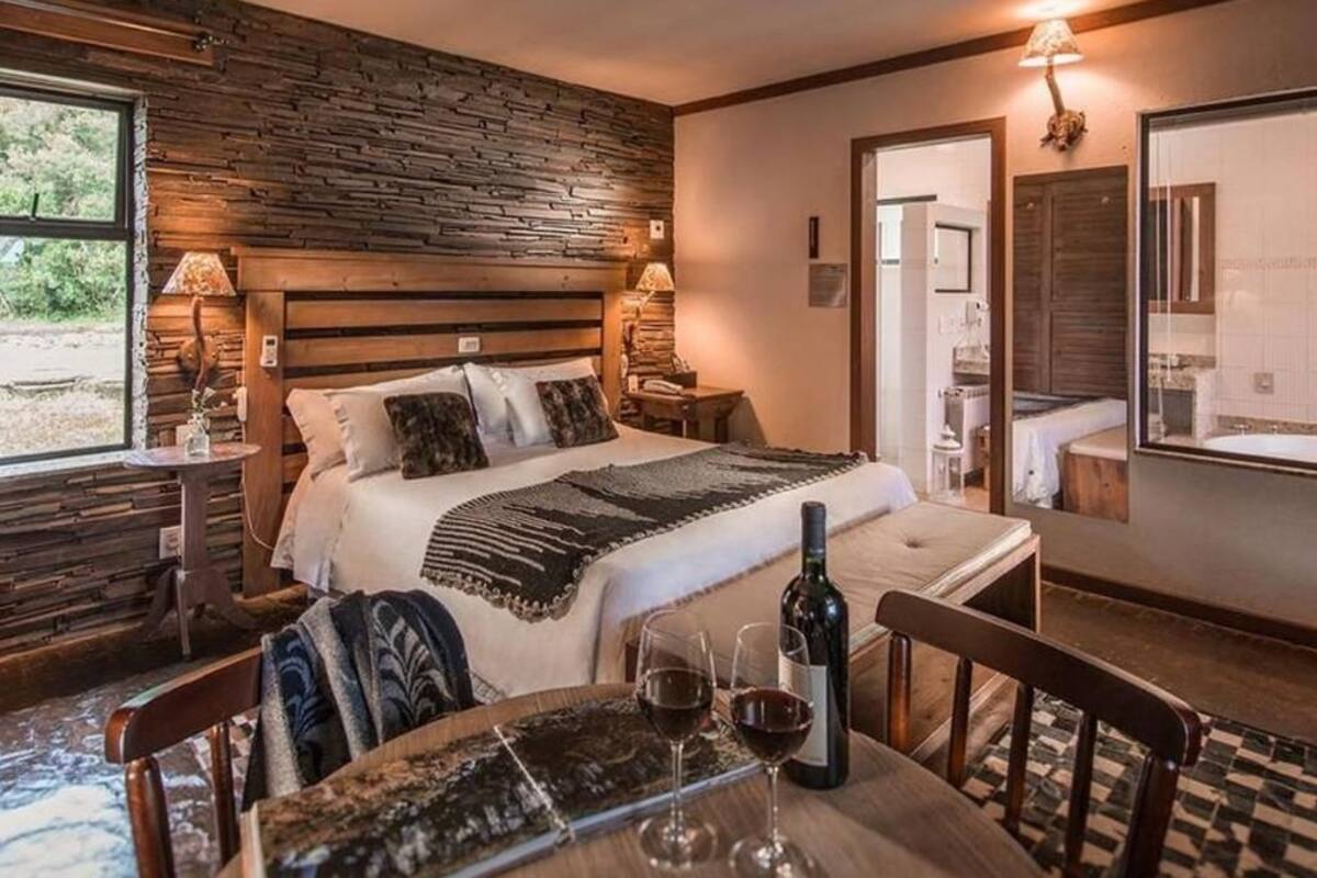 Une chambre à coucher avec une tête de lit en bois, un lit recouvert d’une couette blanche, une table de chevet avec une lampe, et une table à manger avec deux verres à vin et une bouteille.