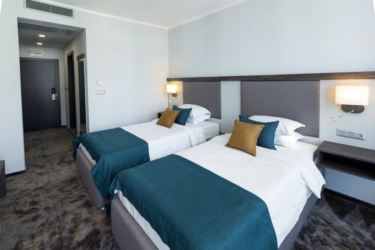 A hotel room with two beds, each with teal and white bedding, a dark headboard, and wall-mounted reading lamps.
