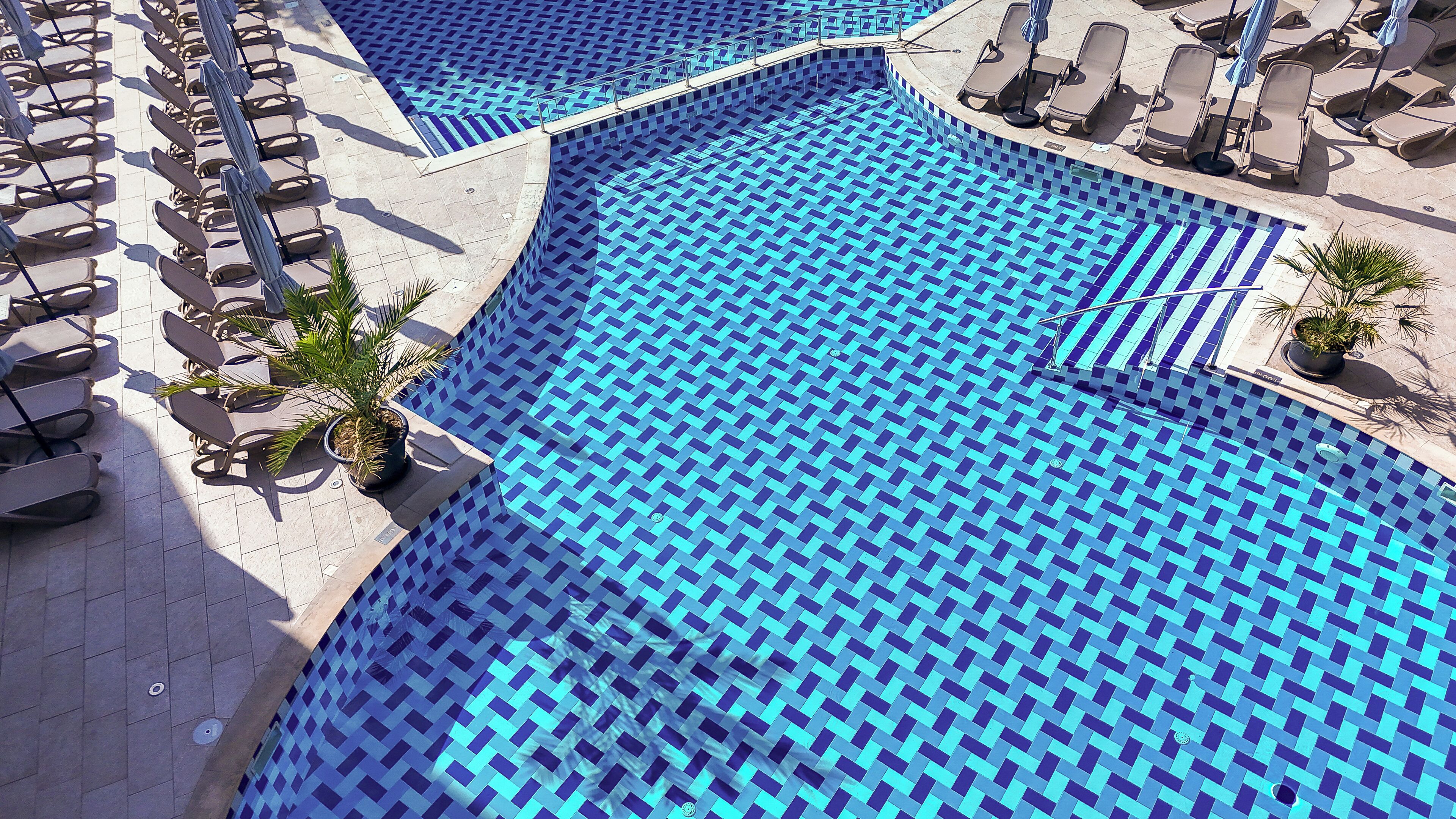 Una amplia piscina de borde infinito con azulejos azules y múltiples sillas y tumbonas alrededor.