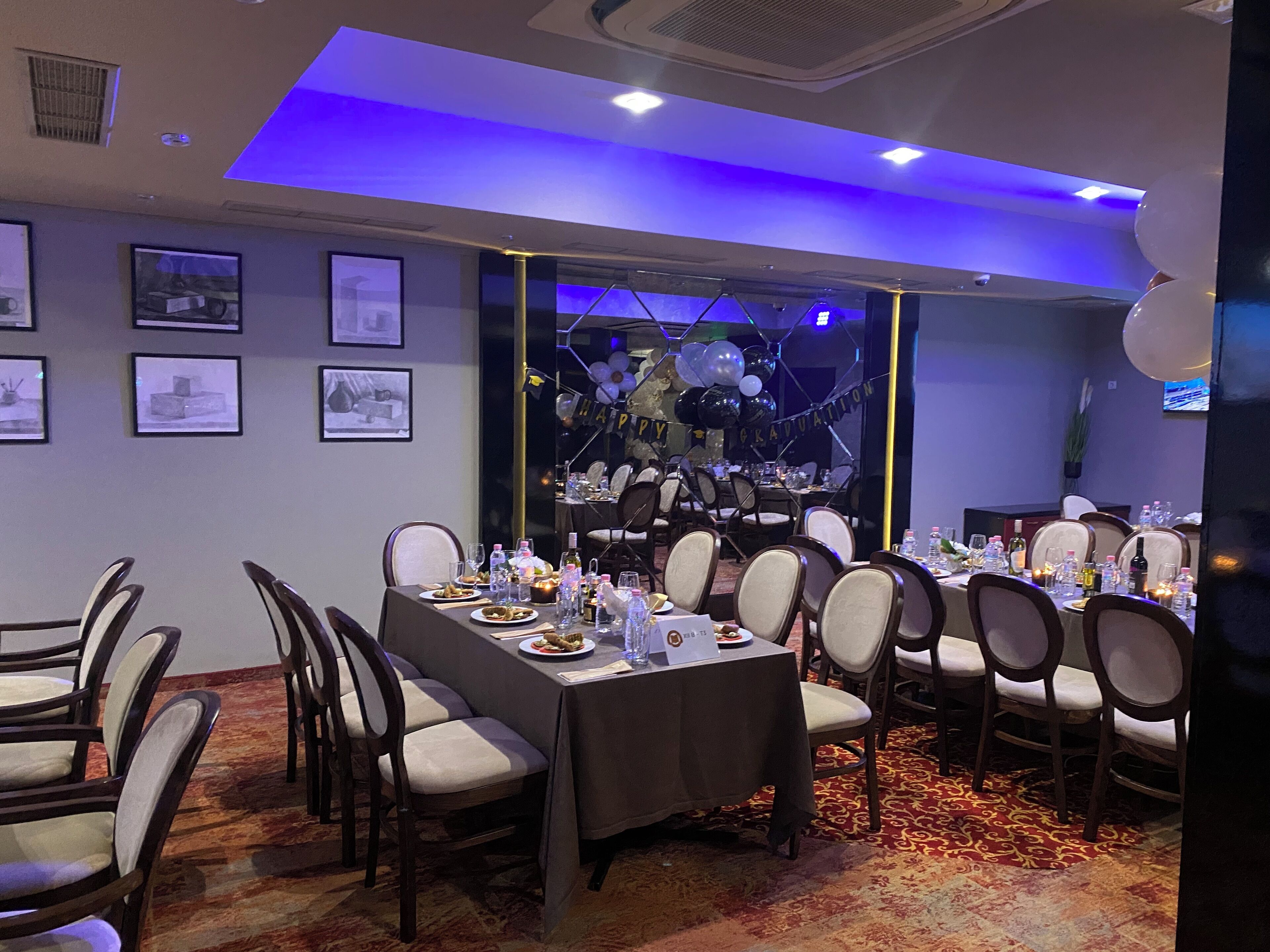 A banquet hall with tables set for a formal event, featuring a ceiling with blue lighting and framed pictures on the walls.