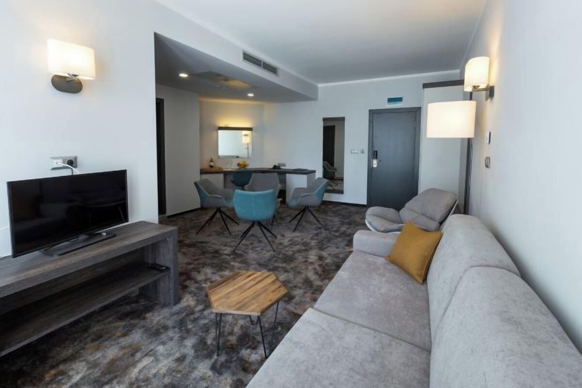 Una habitación de hotel moderna con una televisión de pantalla plana, un sofá gris, una mesa de centro de madera y un área de comedor con sillas azules.