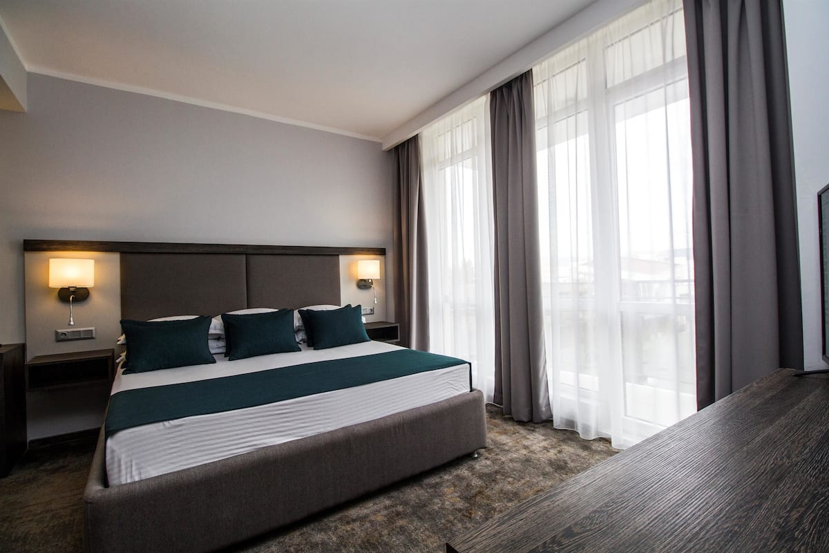 A hotel room with a large bed, two bedside lamps, a wooden headboard, and a view through sheer curtains.