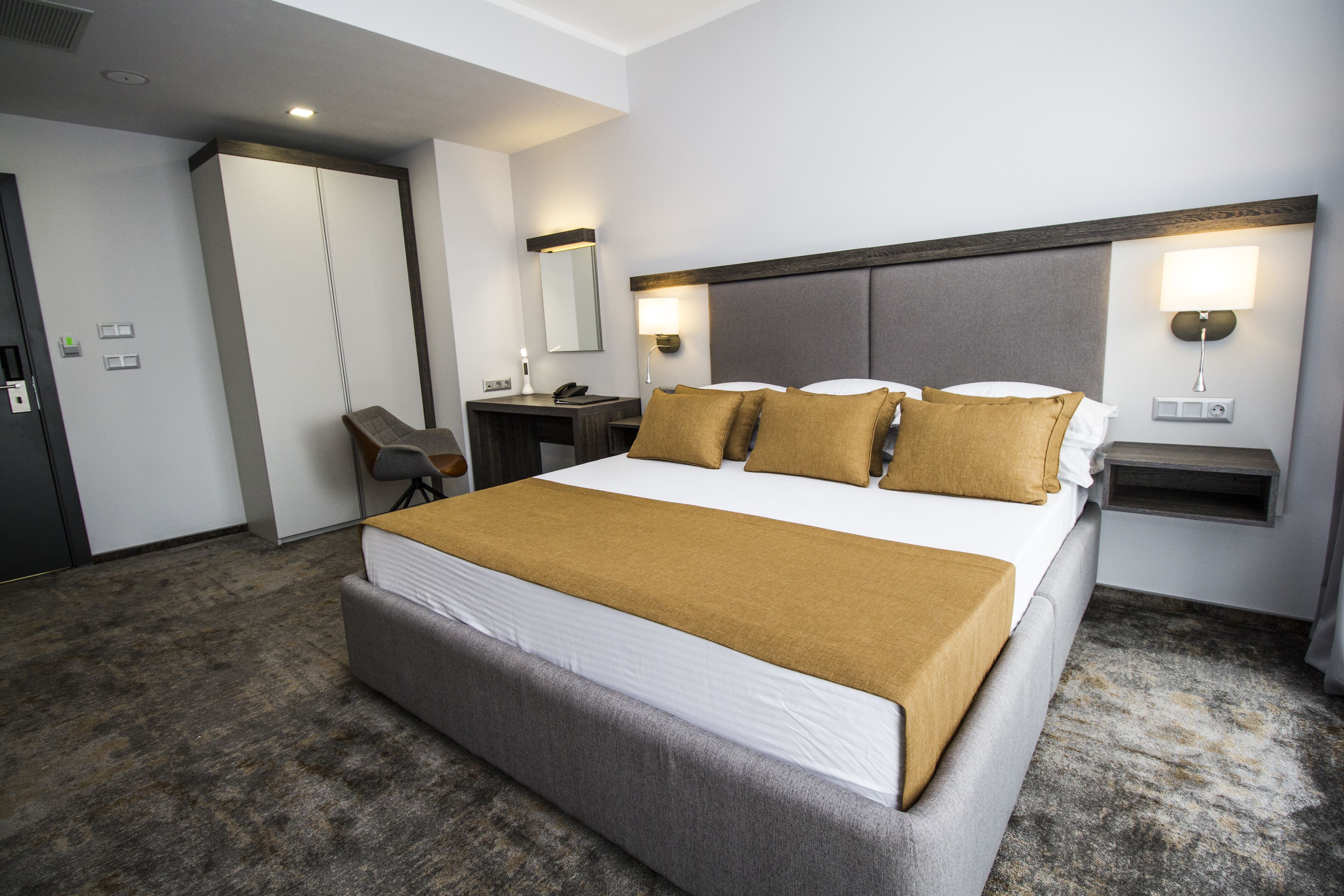 A hotel room with a large bed, a desk, a chair, a wardrobe, and two wall-mounted lamps.