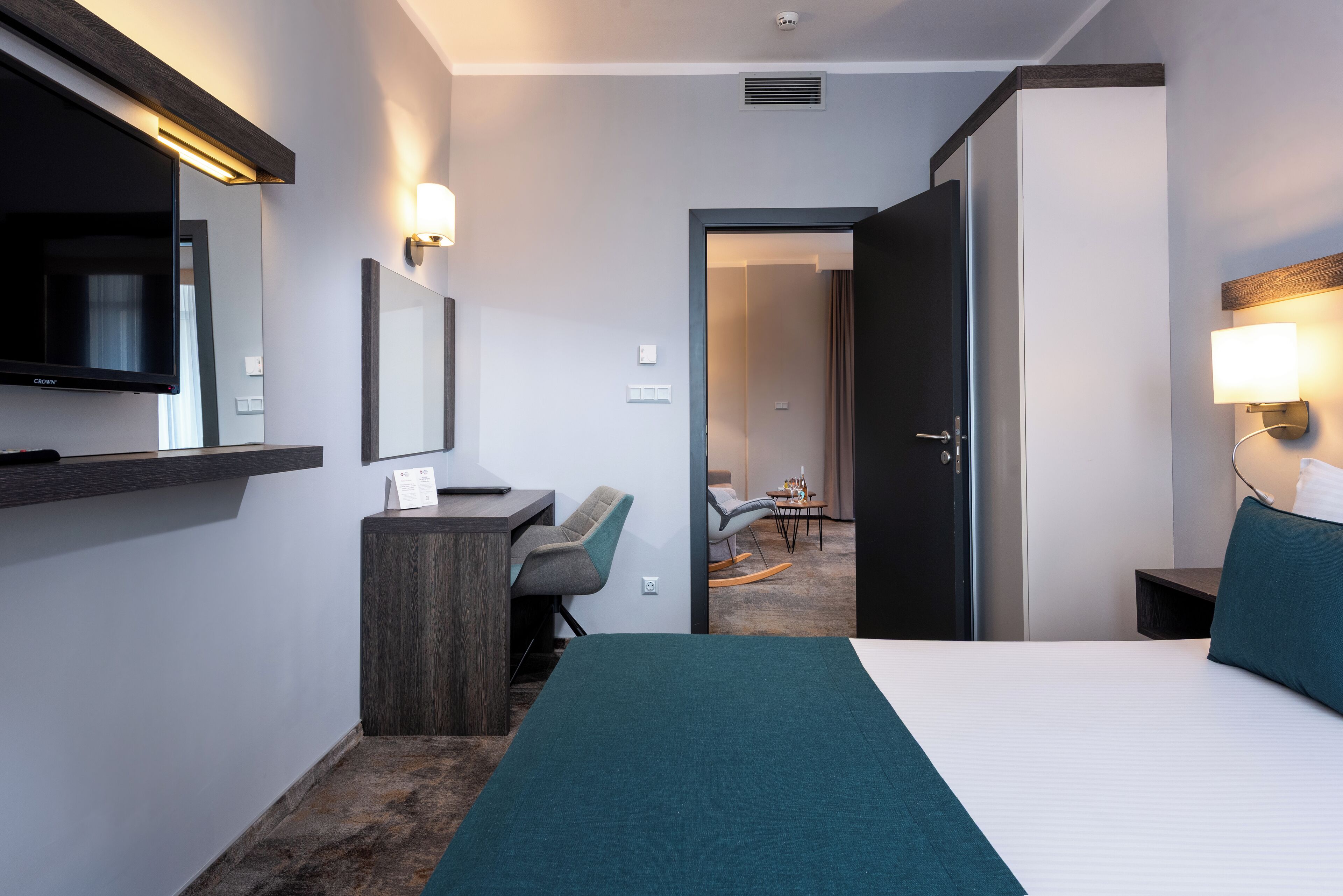 Habitación de hotel con cama, escritorio, silla, televisor y puerta que da a otra habitación.