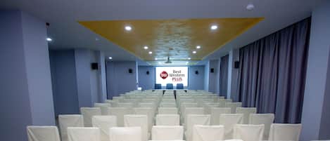 Un salón de conferencias con filas de sillas blancas, un escenario con una pantalla que muestra 'Best Western PLUS', e iluminación empotrada en el techo.