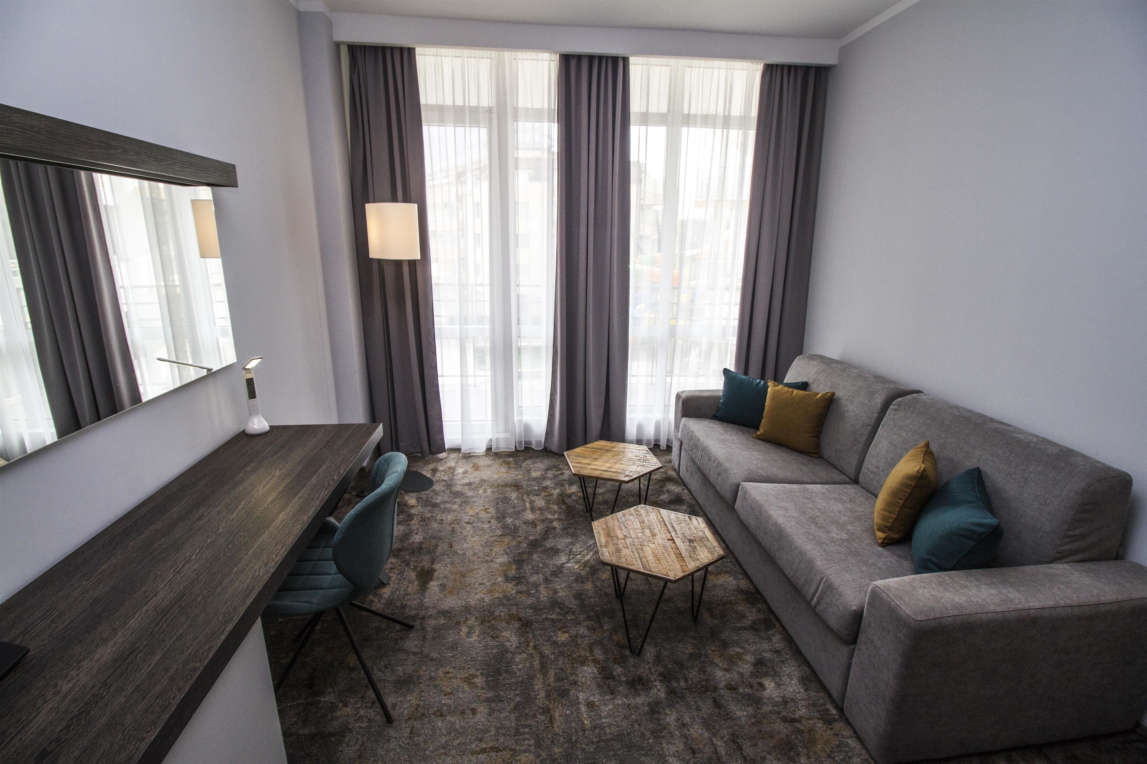 A modern hotel room with a grey sofa, a small wooden coffee table, a desk, and a large mirror.