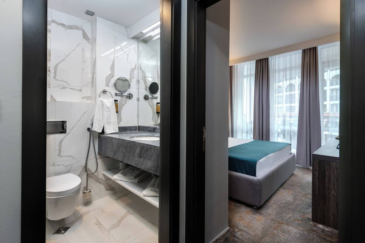 A modern bathroom with a marble vanity, a toilet, and a shower. The room has a bed with a green bedspread and a view of a building through large windows.