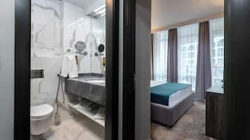 Un baño moderno con tocador de mármol, inodoro y ducha. La habitación tiene una cama con colcha verde y vistas a un edificio a través de amplios ventanales.