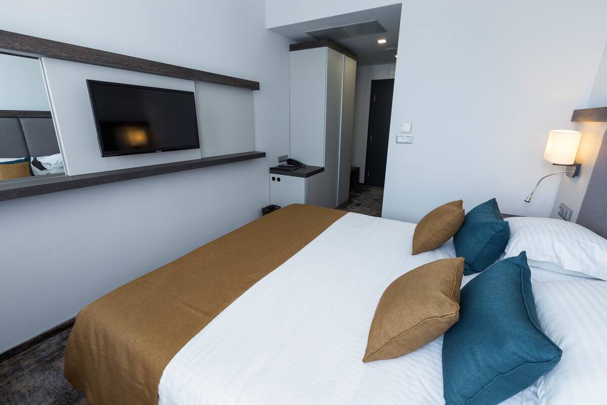 A hotel room with a bed, a TV mounted on the wall, a bedside table, and a lamp.