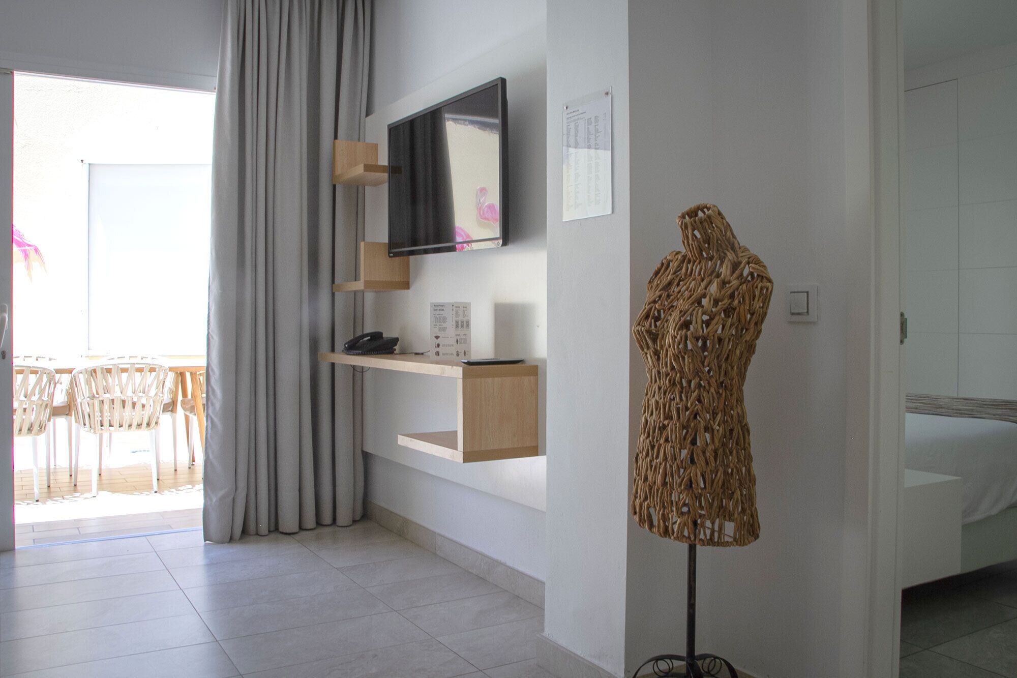 Ein modernes Hotelzimmer mit einem an einer Holzwand montierten Flachbildfernseher, einem Schaufensterpuppenkleid auf einem Ständer und einem Balkon mit Sitzgelegenheiten.