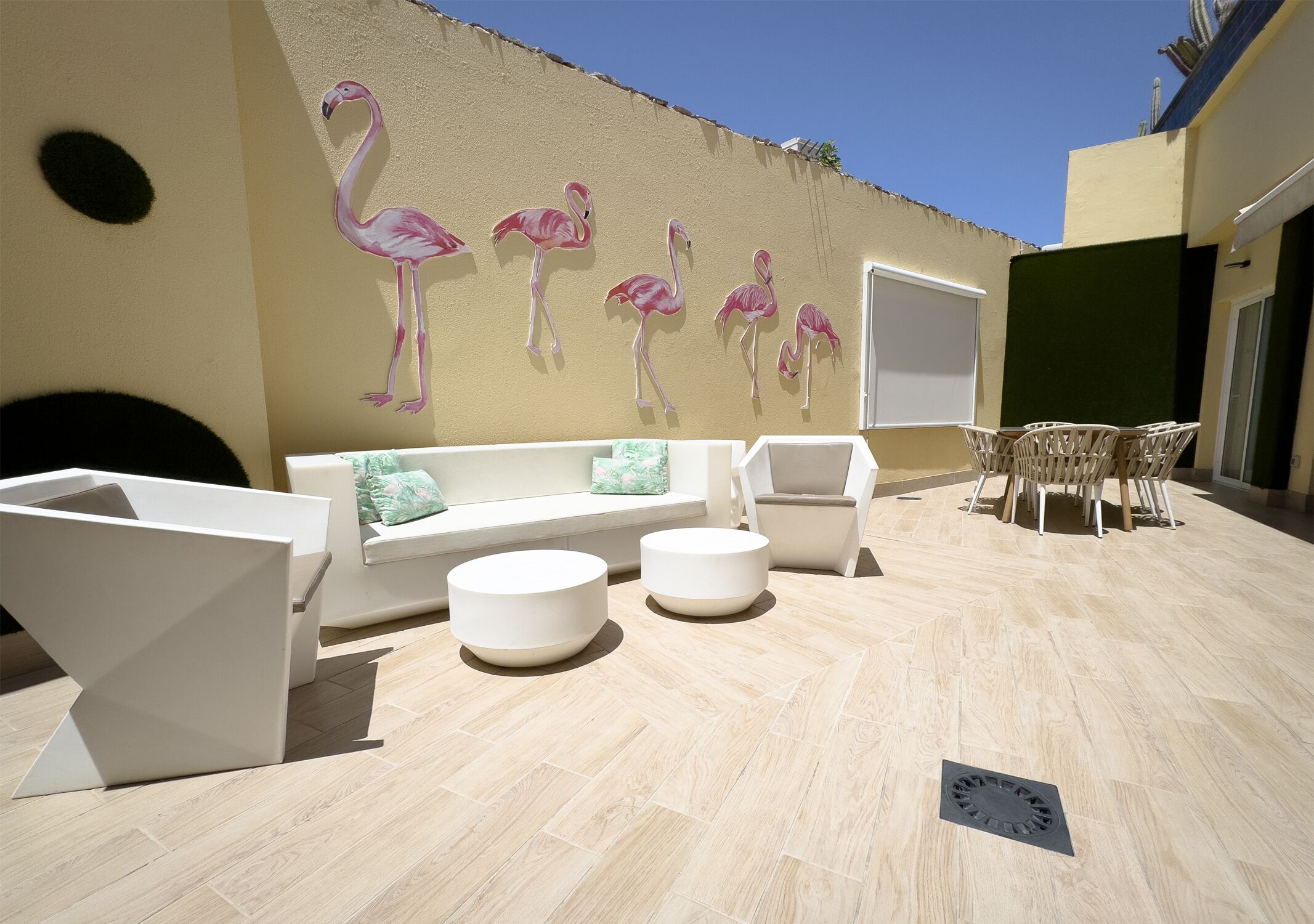Un área de estar al aire libre con sofá, sillas y una mesa, un mural en la pared con flamencos y piso de baldosas.