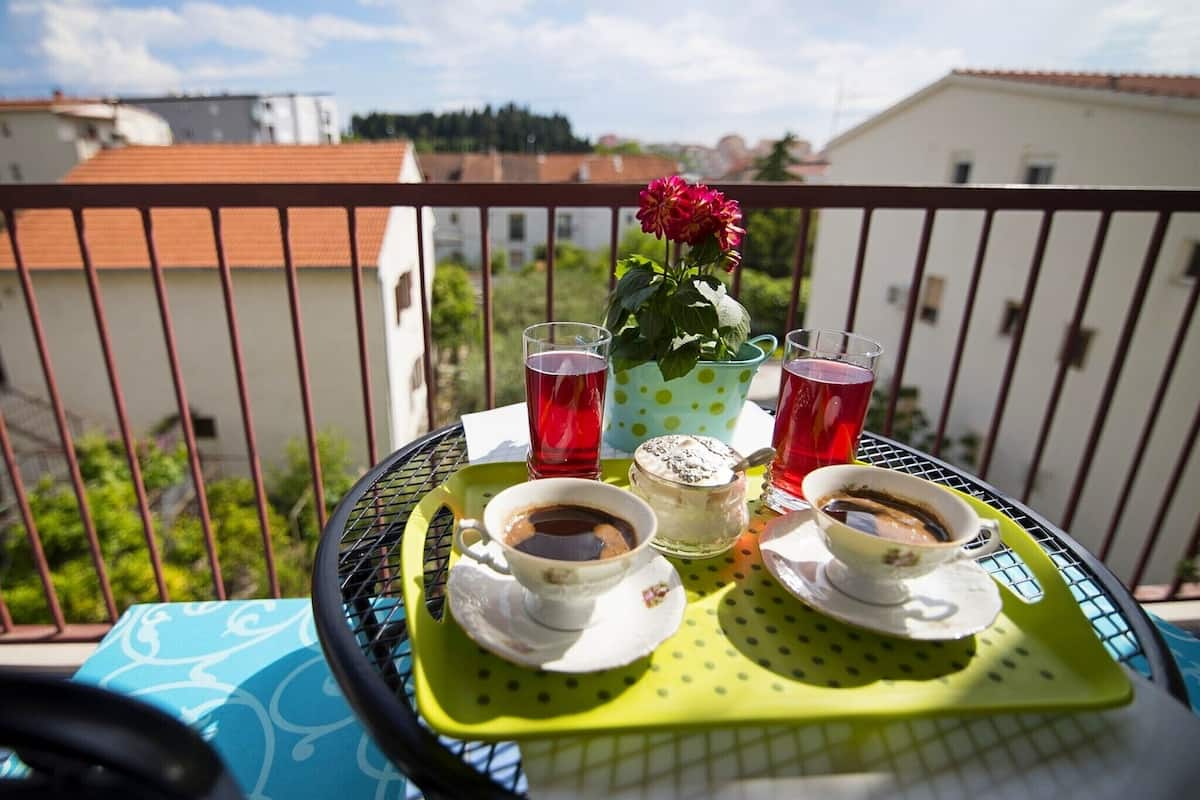 A balcony with a table set for two, featuring two cups of coffee, two glasses of red beverage, and a flower in a polka-dotted pot.