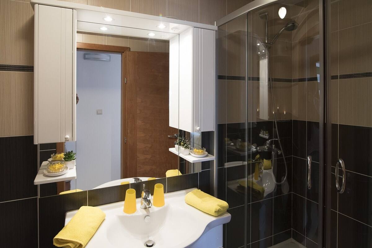 Uma casa de banho moderna com um espelho grande, uma pia com toalhas amarelas e uma área de duche com divisória de vidro.