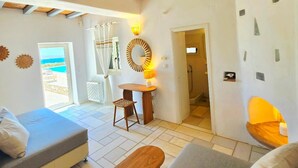 A cozy room with a bed, a wooden bench, and a table with a lamp. There is a view of the sea through a door. - Paros Afrodite Luxury Villas (Paros)