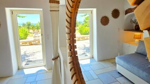 A modern interior with a wooden spiral staircase, a grey sofa, and a view of a garden through a glass door. - Paros Afrodite Luxury Villas (Paros)