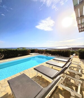 En pool med solstolar, en stenbelagd uteplats och utsikt över havet och de avlägsna bergen.