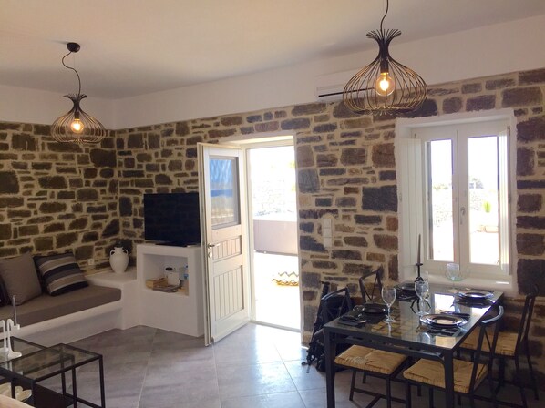 A living room with a stone wall, a TV, a sofa, a glass coffee table, and a dining area with a table set for two.