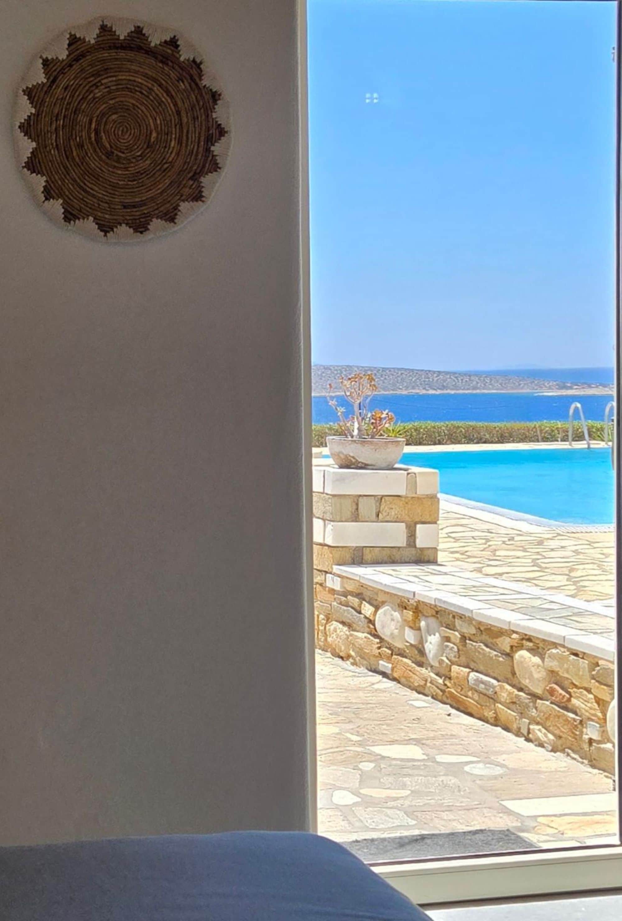 A view from a room with a stone wall and a large window overlooking a pool and the sea.