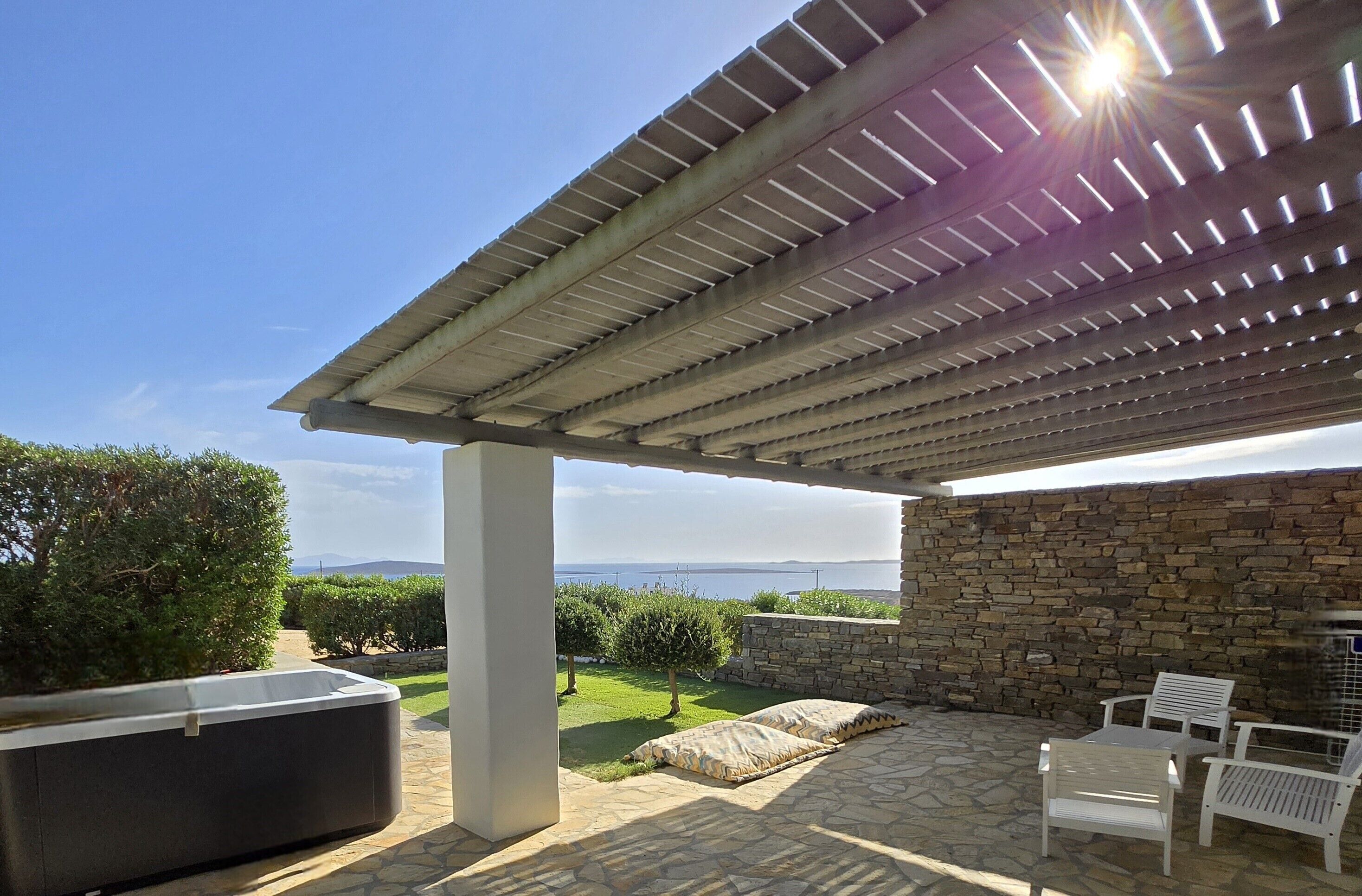 An outdoor patio with a stone wall, a pergola, a hot tub, and white chairs.
