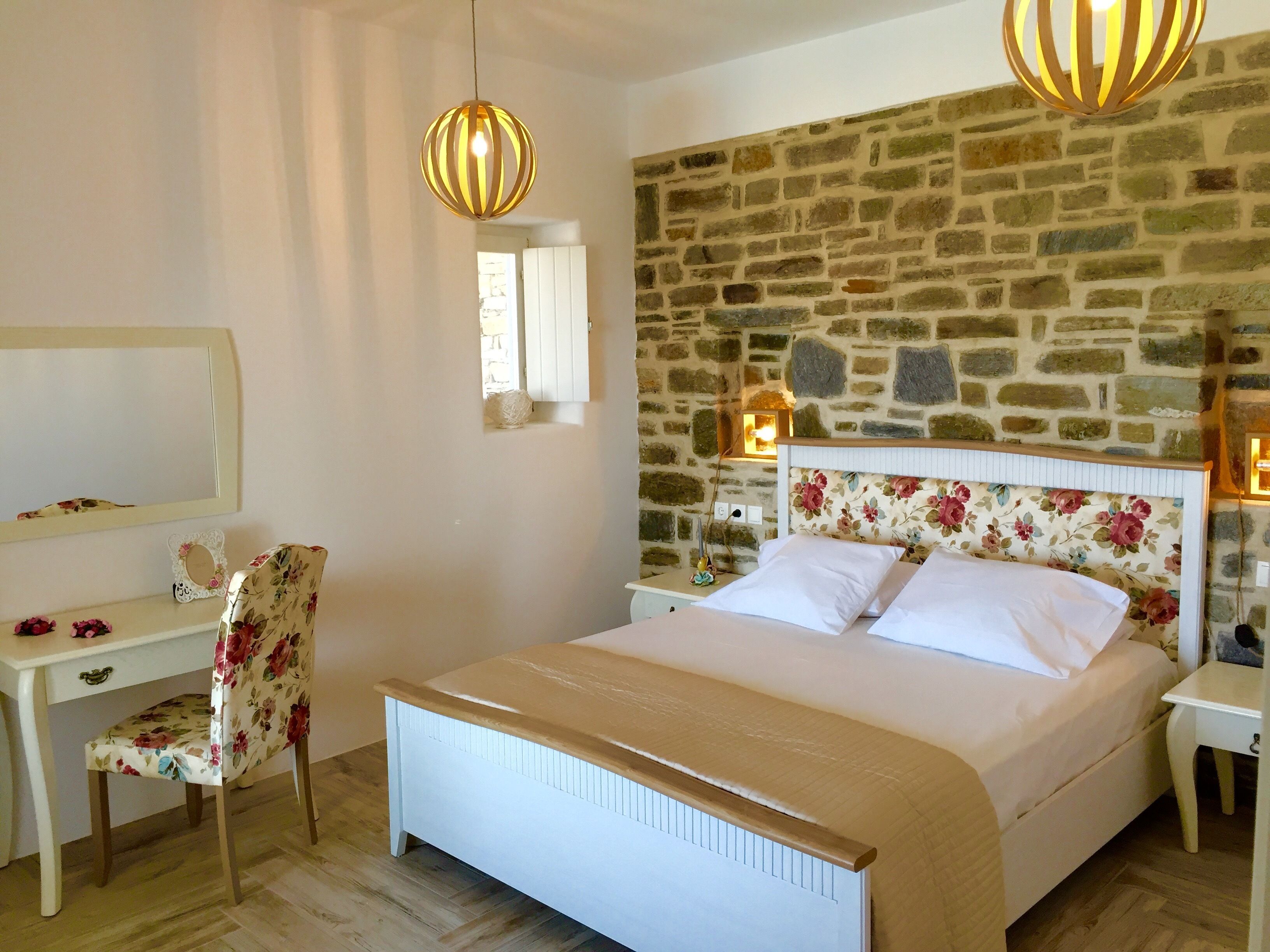 A bedroom with a stone wall, a bed with floral headboard, a desk, and two pendant lights.