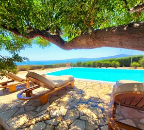 A poolside area with lounge chairs, a stone-tiled floor, and a view of the sea and mountains.