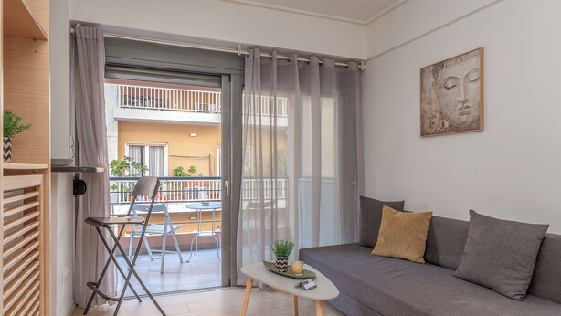 A modern living room with a grey sofa, a small white table, and a balcony with a chair and plants. - Elvita Apartments (Athens)