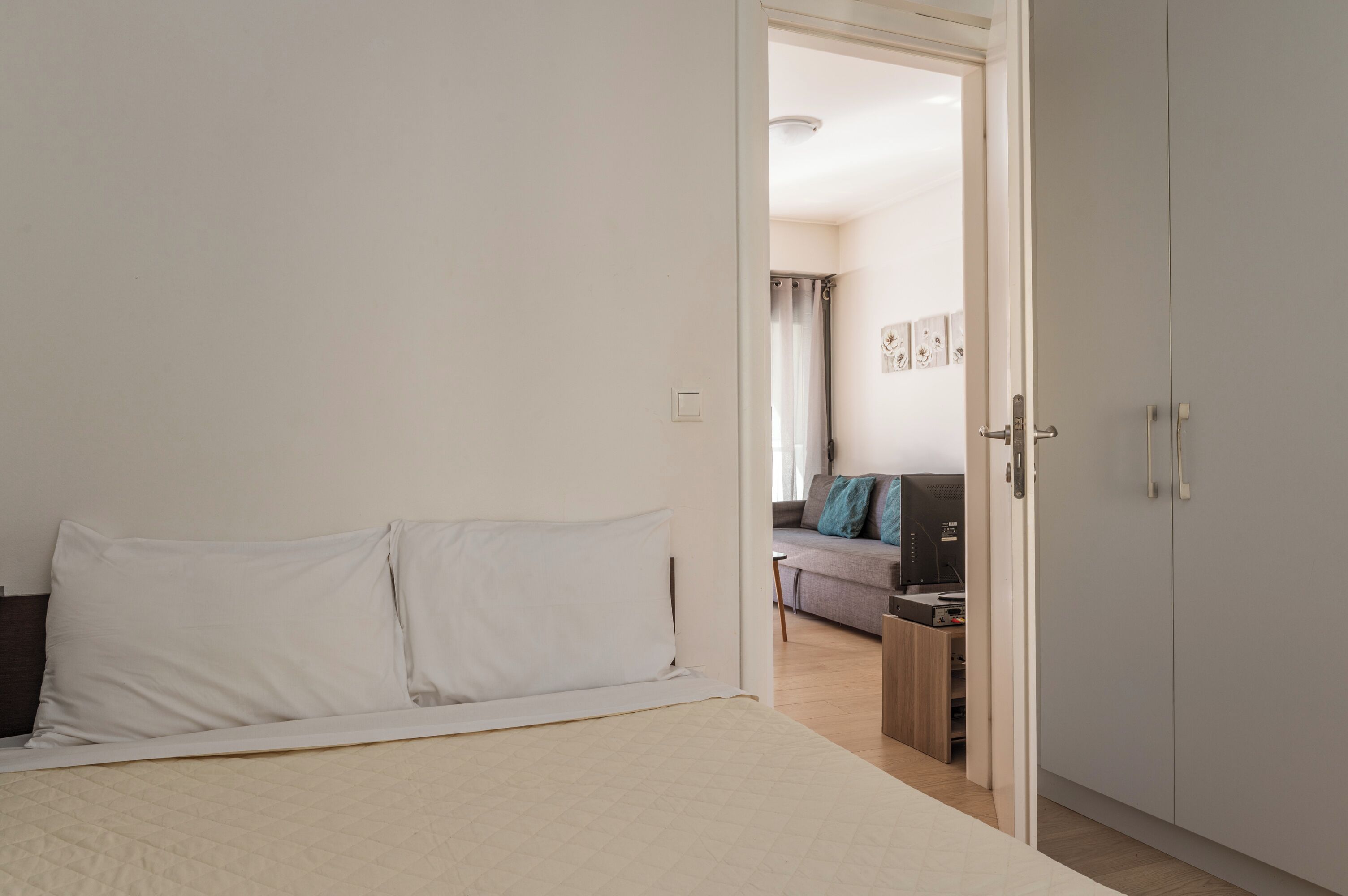 A bedroom with a bed, a wardrobe, and a view into a living area with a sofa and a desk.