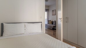 A bedroom with a bed, a wardrobe, and a view into a living area with a sofa and a painting on the wall. - Elvita Apartments (Athens)