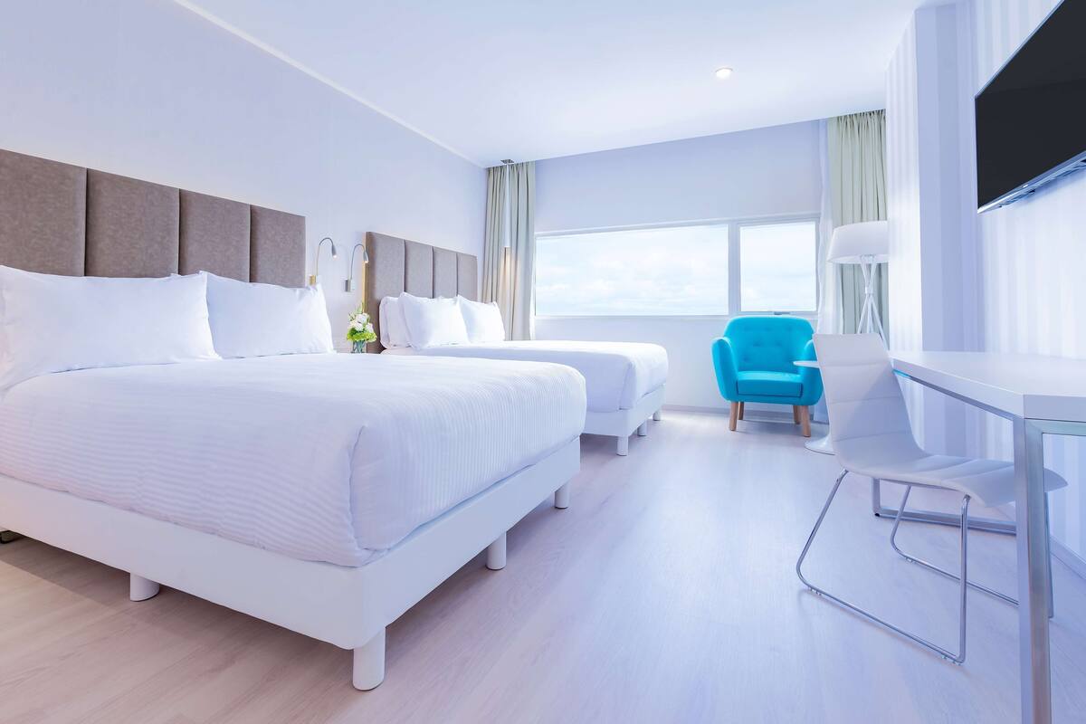 A hotel room with two beds, a desk, a chair, a TV, and a large window.