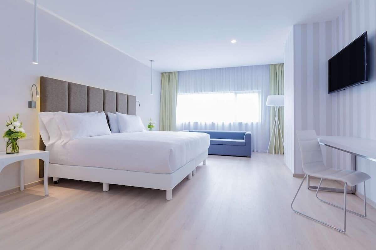 A modern hotel room with a bed, a desk, a chair, a TV, and a window with curtains.