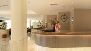 A hotel reception area with a marble floor, a reception desk, potted plants, and a wall clock. - Hotel Garbí Cala Millor (Sant Llorenc des Cardassar)