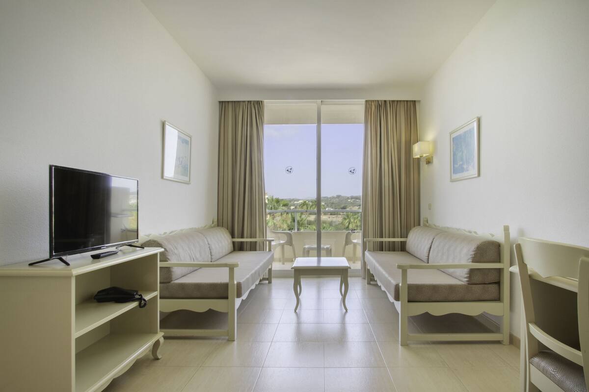 A hotel room with a TV, two sofas, a small table, and a balcony with curtains.
