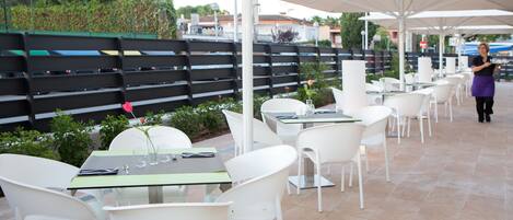 An outdoor dining area with white tables and chairs, large umbrellas, and a black fence in the background.