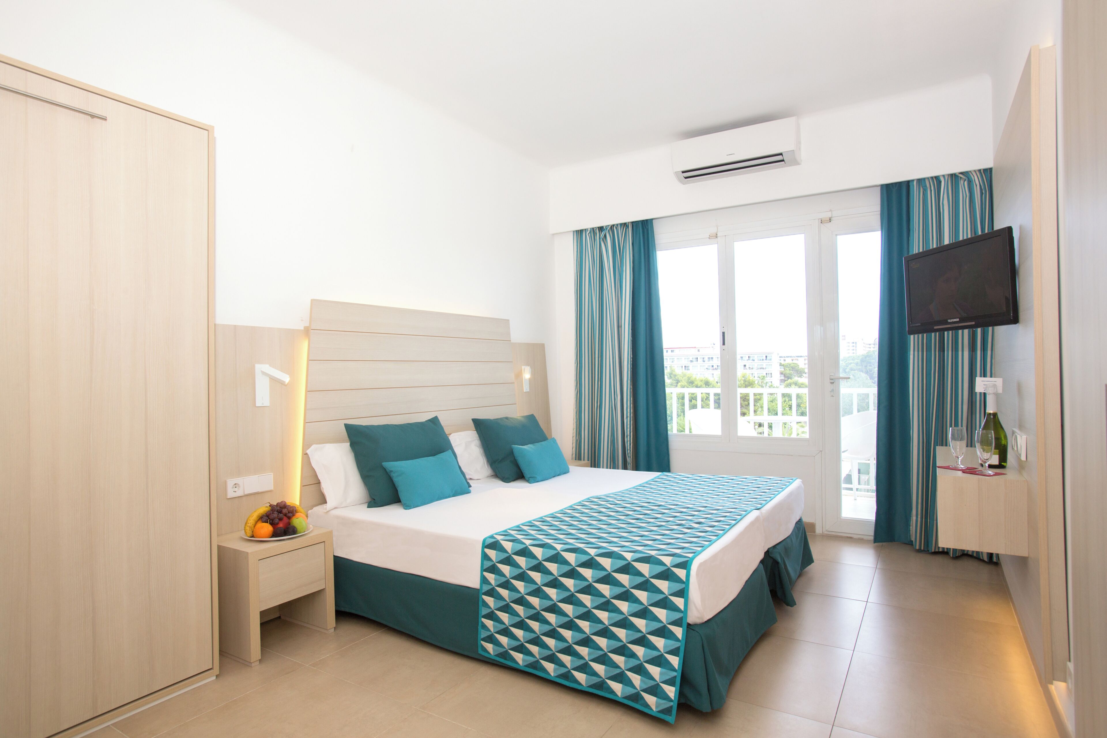 A hotel room with a bed, bedside table, fruit bowl, curtains, and a TV.