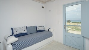 A white room with a blue door, a white sofa with striped and polka-dotted cushions, and a view of a beach and mountains through the door. - Depis Bay Apartments (Naxos)